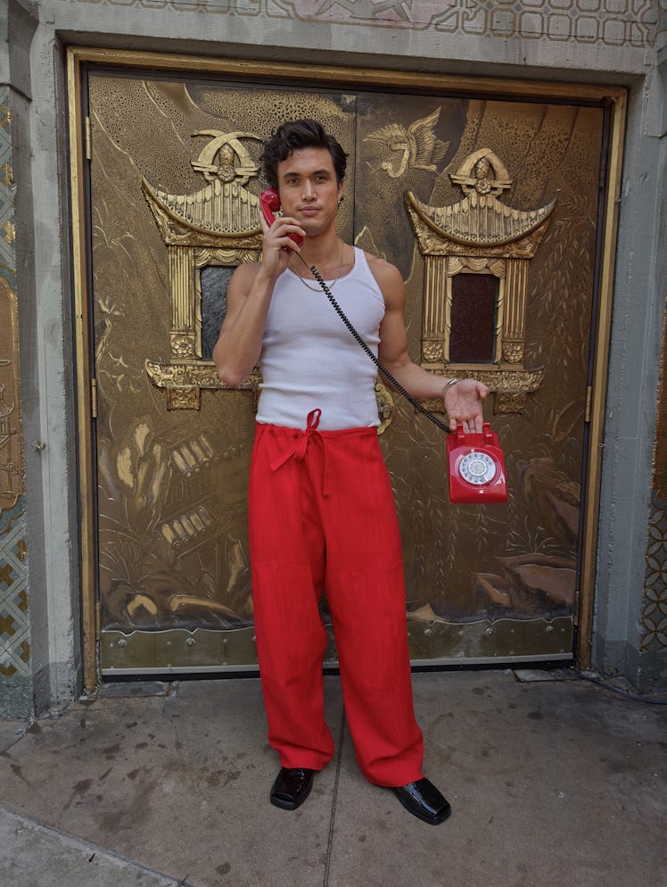 Charles Melton wears a white tank top, red pants, watch, gold necklace and black shoes.