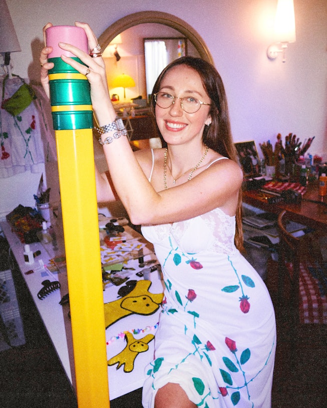 Artist Keely Murphy leans on a table holding a human-sized yellow pencil, and in the background, you...