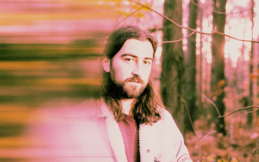 A man with a beard is standing in a forest during autumn, with a blurred pink and yellow light effec...