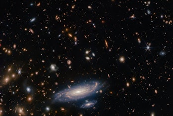 photo of a spiral galaxy in the foreground, with hundreds of tiny galaxies of different shapes and c...