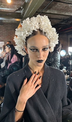 A model wearing a white flower crown and back graphic, gothic eyeliner backstage at Rodarte's FW 23 ...