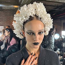 A model wearing a white flower crown and back graphic, gothic eyeliner backstage at Rodarte's FW 23 ...