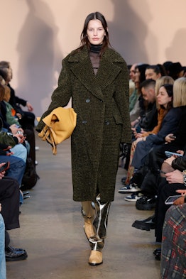 a model walks a runway wearing a big coat and a tote bag