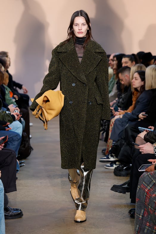 a model walks a runway wearing a big coat and a tote bag