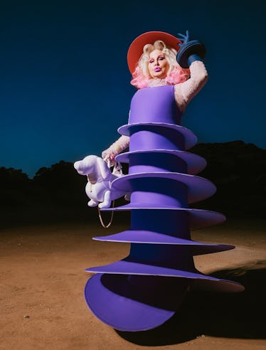 Jennifer Coolidge wears a purple custom design dress, pink straw hat, dog design hatbag and boots.