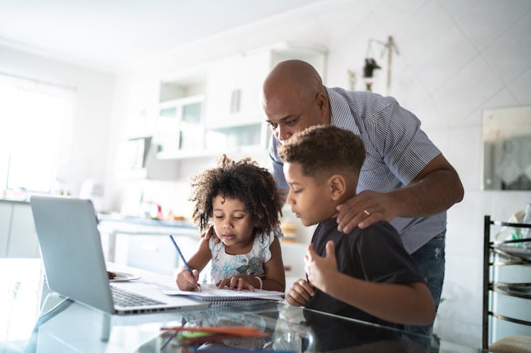 A dad with his hands on his kids' shoulders while they are studying at a table.