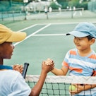 Two kids shake hands over a tennis net.