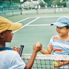 Two kids shake hands over a tennis net.