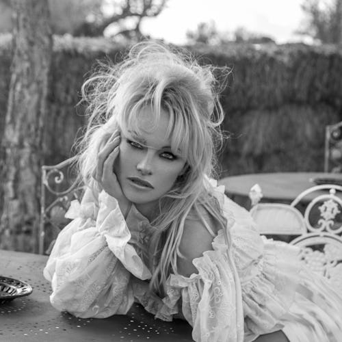 Pamela Anderson wears a white dress and sits at a table.