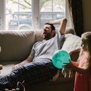 A man on a couch yawning as his two kids play around him.