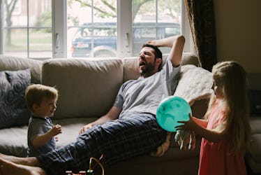 A man on a couch yawning as his two kids play around him.