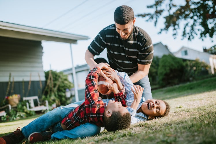 A dad playing football and wrestling with his two kids outside on grass.