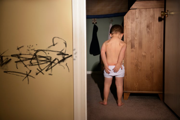 A boy in underwear holding his butt in pain, beside a wall with black scribbles on it.
