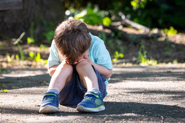 A child sits outside on the ground with his head in his hands.
