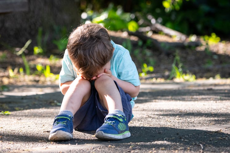 A child sits outside on the ground with his head in his hands.