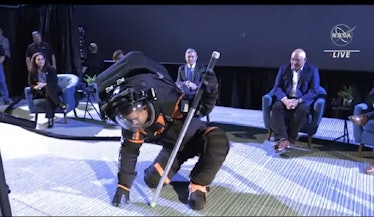 a man in a black and orange spacesuit bends over to touch the ground