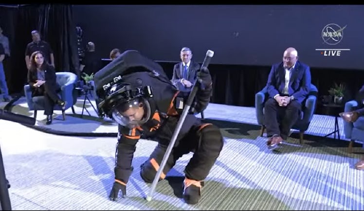 a man in a black and orange spacesuit bends over to touch the ground