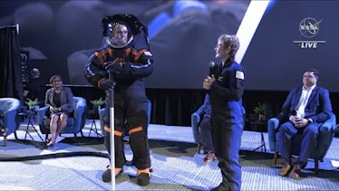 A female astronaut in a blue flightsuit stands next to a man in a black and orange spacesuit holding...