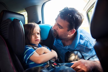 A child crosses their arms and looks grumpy, refusing to get out of their car seat, as their dad lea...