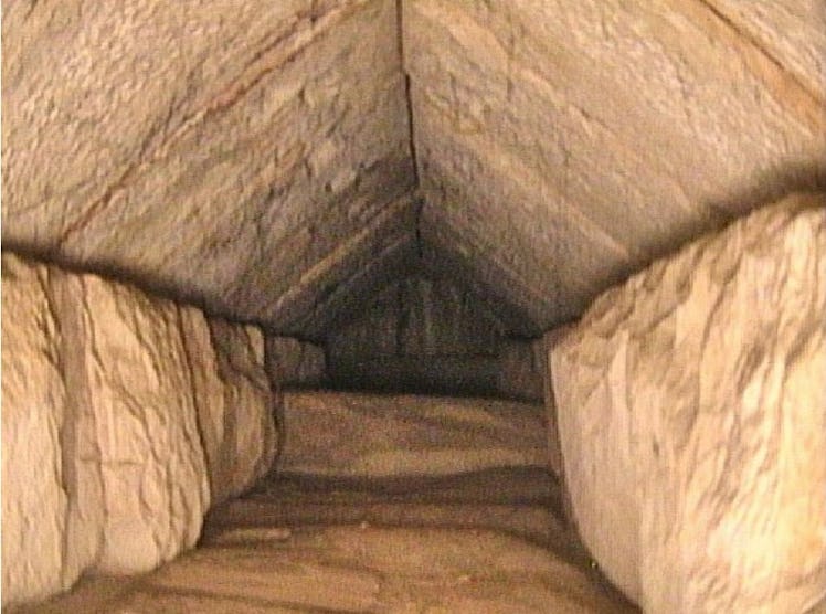 A look down a straight tunnel with a peaked ceiling. The ceiling, floor, and walls are of brown rock...