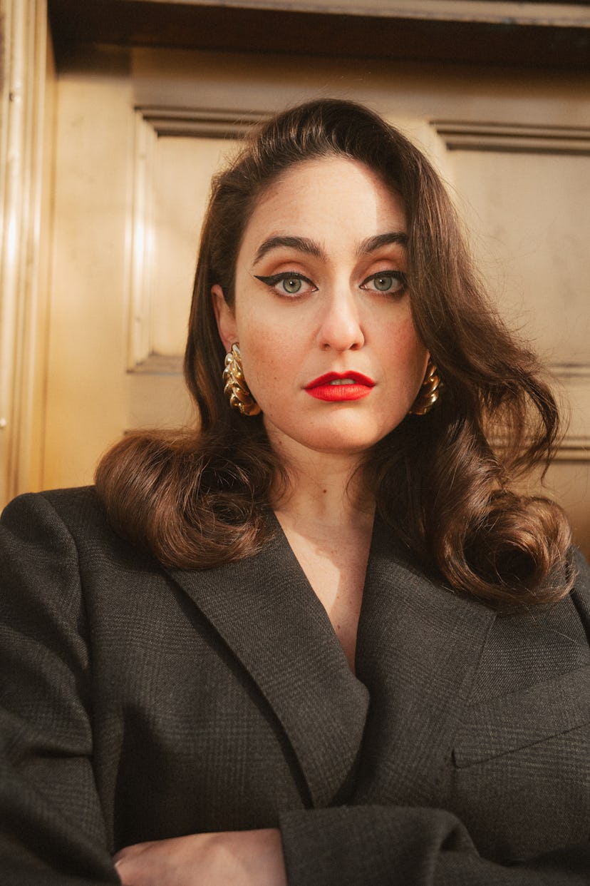 Comedian Catherine Cohen wearing a blazer, red lipstick and gold earrings.