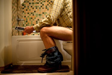 A man sitting on the toilet with his jeans at his ankles, reading a magazine.