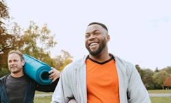Two men smiling and holding rolled up yoga mats after exercising outdoors.