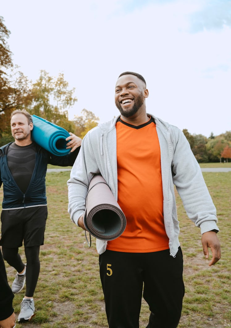 Two men smiling and holding rolled up yoga mats after exercising outdoors.