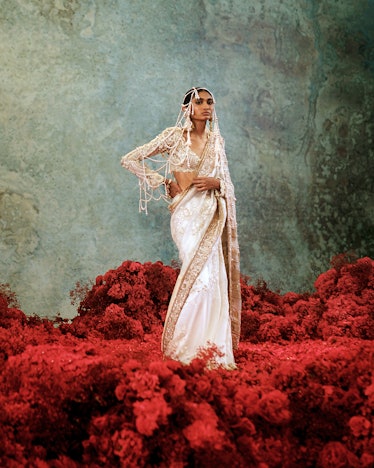 a model standing among red flowers wearing an intricate Indian bridal ensemble. 