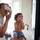 A dad and son in the bathroom putting shaving cream on their faces in front of a mirror.