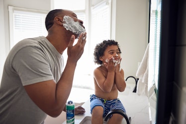 A dad and son in the bathroom putting shaving cream on their faces in front of a mirror.
