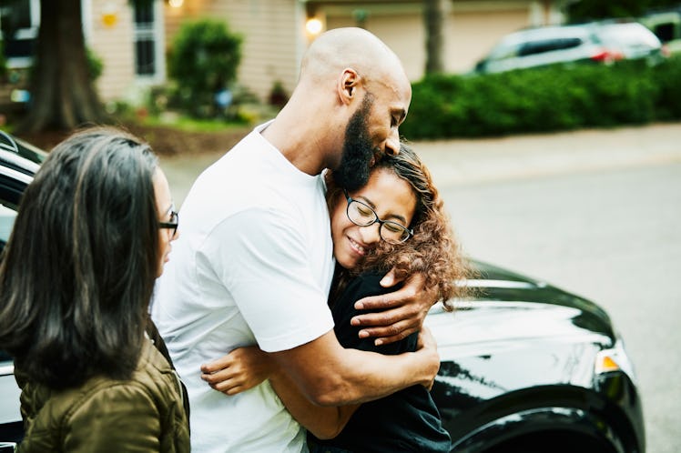 A father embracing his trans teenager, standing beside the kid's mother, in front of a car outside.