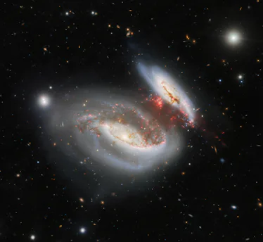 two spiral galaxies in the center of a black background, with streamers of red gas stretching betwee...