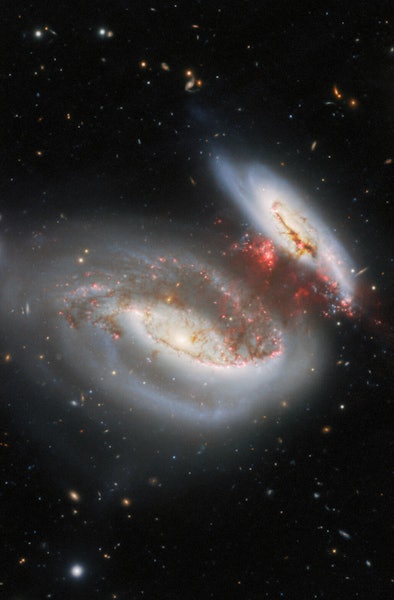 two spiral galaxies in the center of a black background, with streamers of red gas stretching betwee...