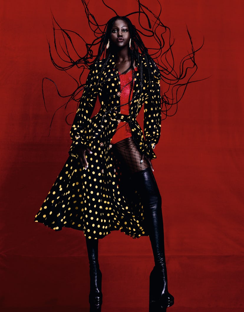 the model adut akech posing against a red background wearing a black trench coat with yellow polkado...