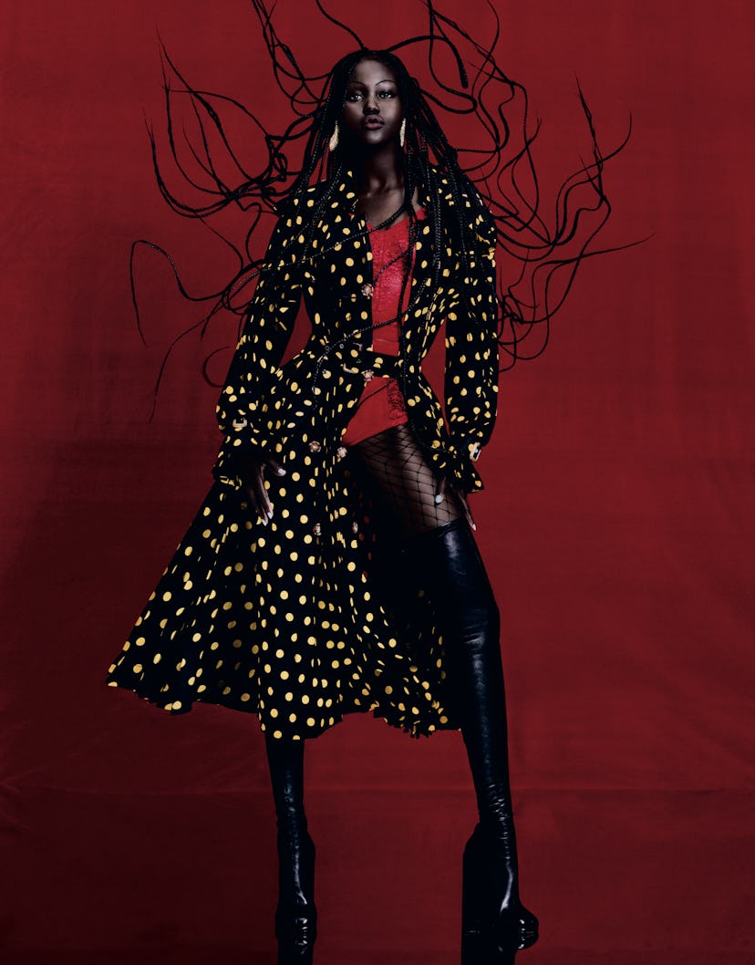 the model adut akech posing against a red background wearing a black trench coat with yellow polkado...