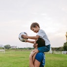 A child sitting on their dad's shoulders, holding a soccer ball, as dad walks across a soccer field.