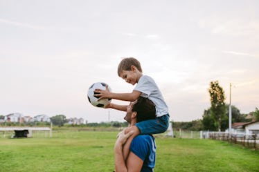 A child sitting on their dad's shoulders, holding a soccer ball, as dad walks across a soccer field.