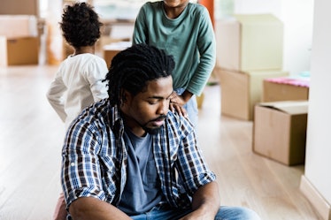 A divorced man sitting on the floor, upset, while his kids and packed boxes in an empty room are beh...