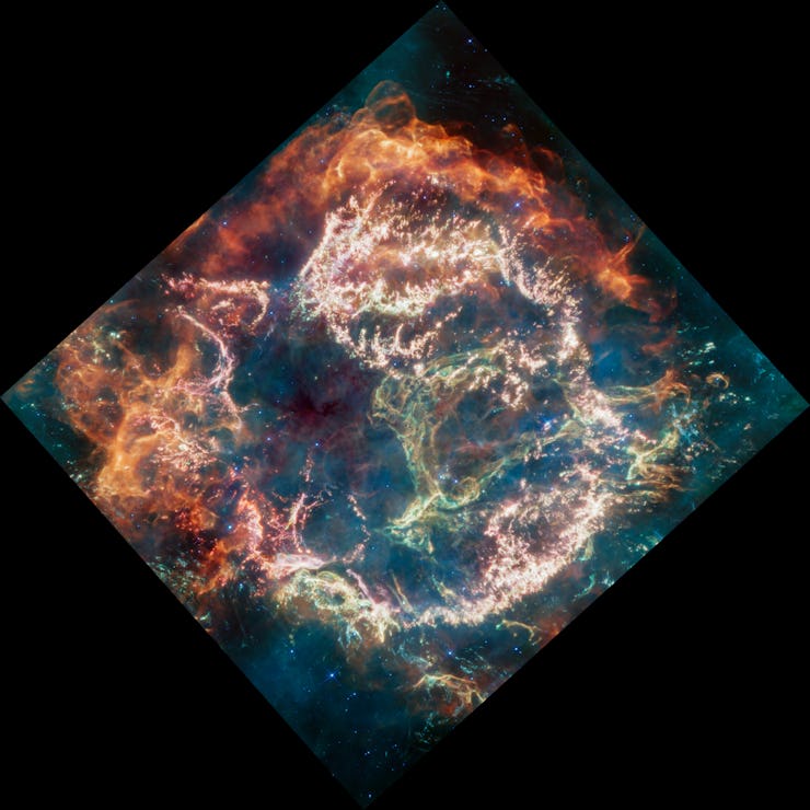 Filaments and clumps of gas and dust expand outward in a roughly circular pattern, in colors ranging...