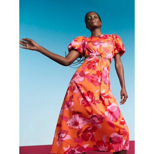 A model poses confidently in a vibrant orange floral dress with puffed sleeves, set against a blue g...