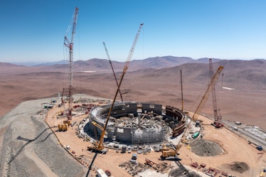 a fleet of cranes surrounding a round, partially completed building at a desert construction site