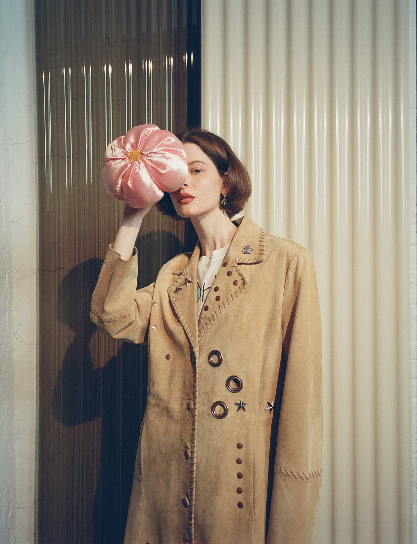 a model wearing a camel-colored suede jacket embellished with metal studs and rivets holds a puff of...