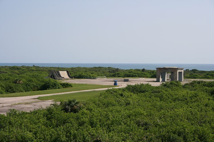 photo of a gray concrete pad with a concrete structure on it amid green vegetation with blue water v...