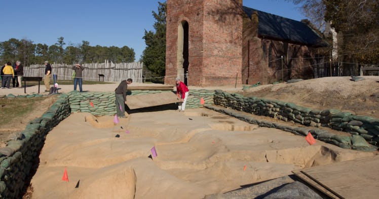 green sandbags line a square sandy excavation pit with a brick building and blue sky in the backgrou...