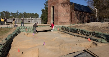 green sandbags line a square sandy excavation pit with a brick building and blue sky in the backgrou...