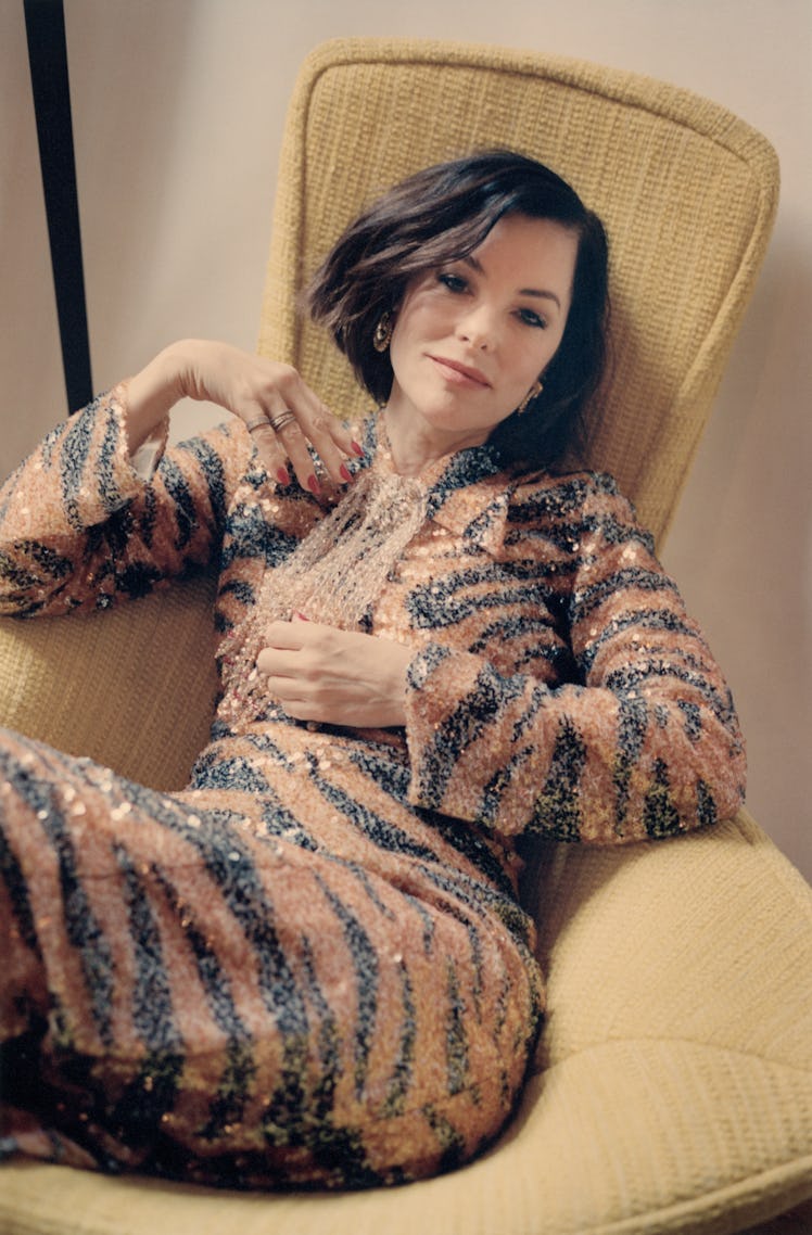 Parker Posey wearing a tiger print, sequined Gucci dress, lounges in a mid-century yellow chair