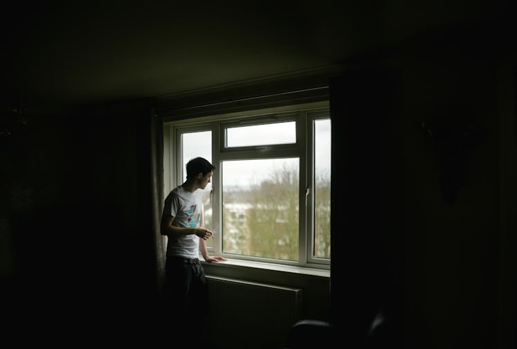 A man in a dark room, staring out a window, smoking a cigarette.