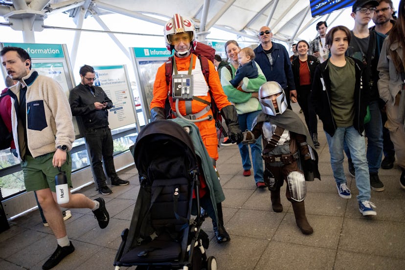 A fan dressed as an X-wing pilot pushes a stroller while holding the hand of a child dressed as a Ma...