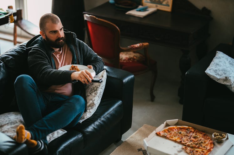 A man watching TV on the couch, eating pizza and drinking beer.